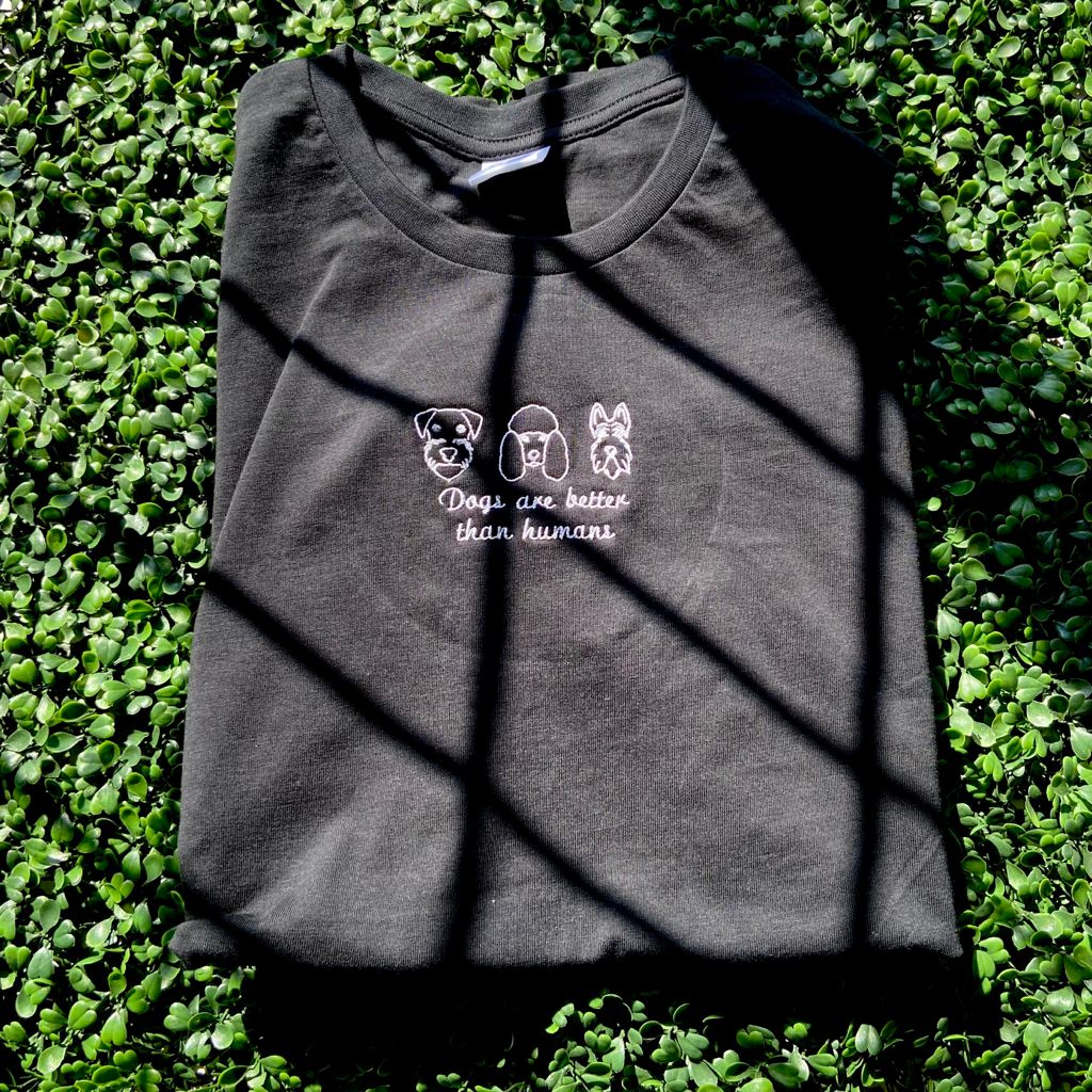 Folded black t-shirt with embroidered dog faces and 'Dogs are better than humans' text on greenery
