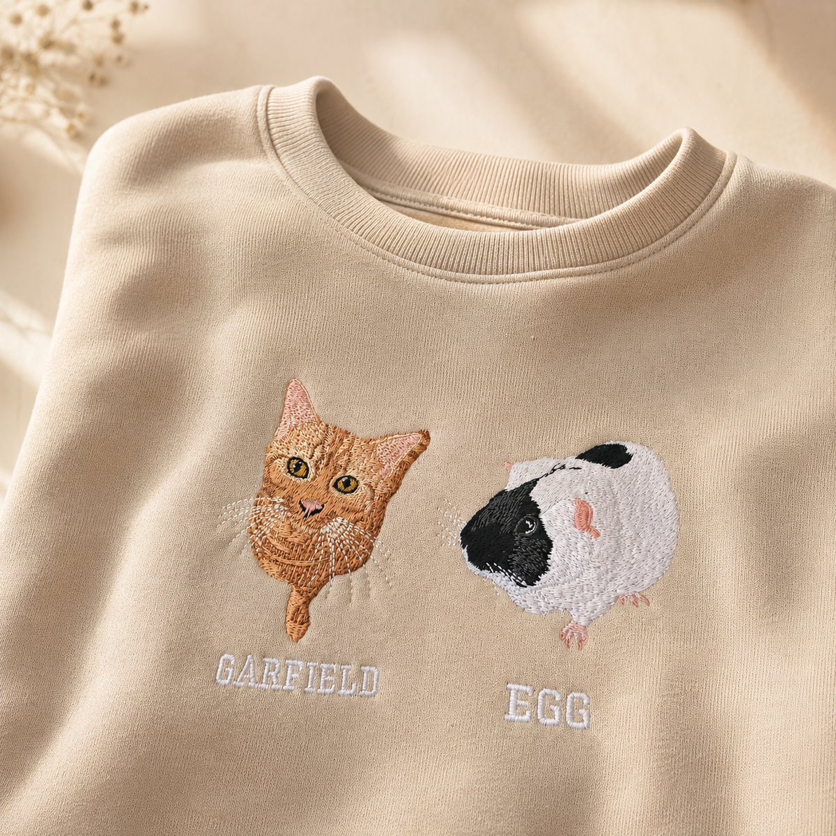 Close-up of bespoke embroidered pet portraits Garfield the cat and Egg the guinea pig on a beige personalised sweatshirt